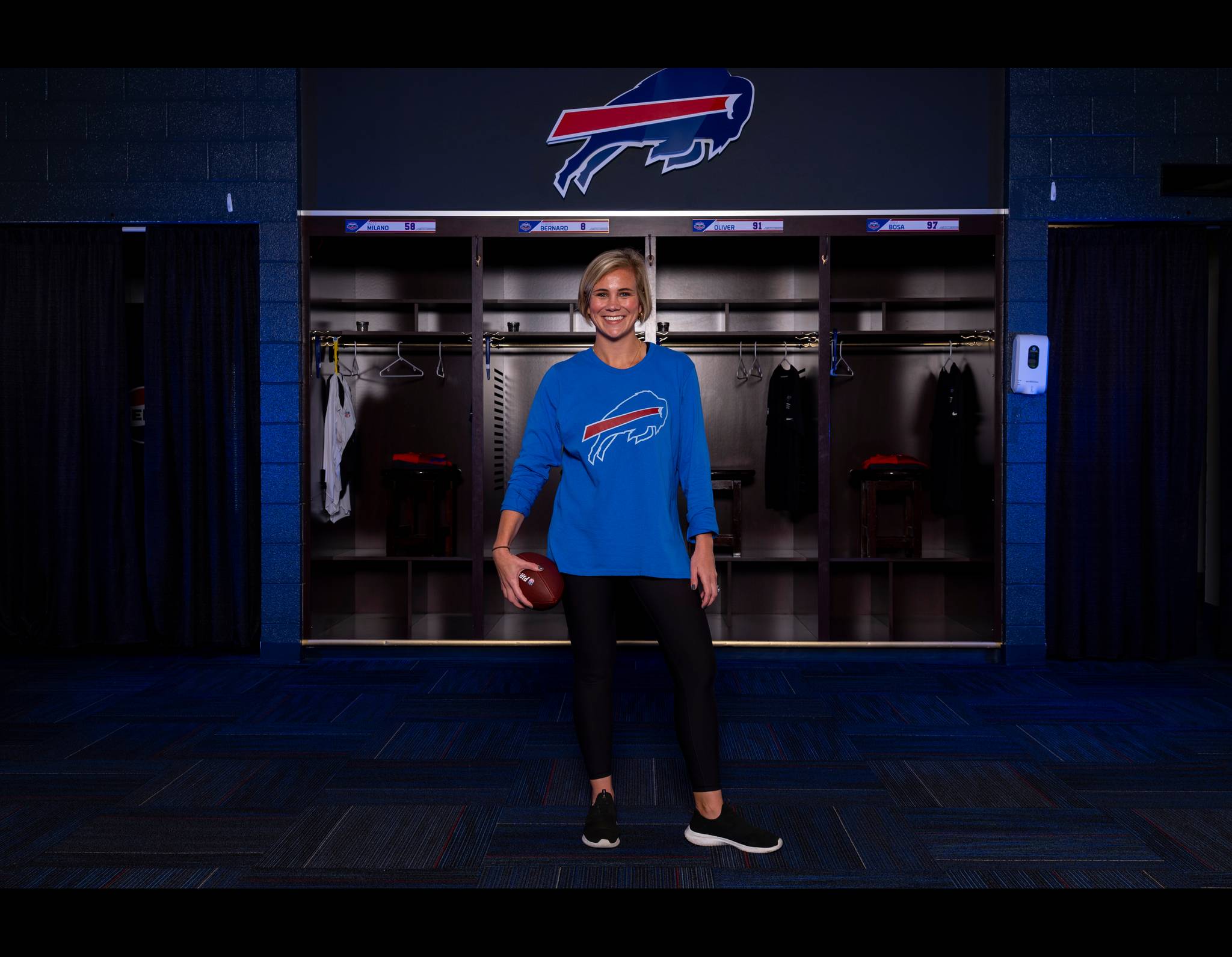 Smiling person in blue Bills shirt holding football, standing in team locker room with player lockers behind her