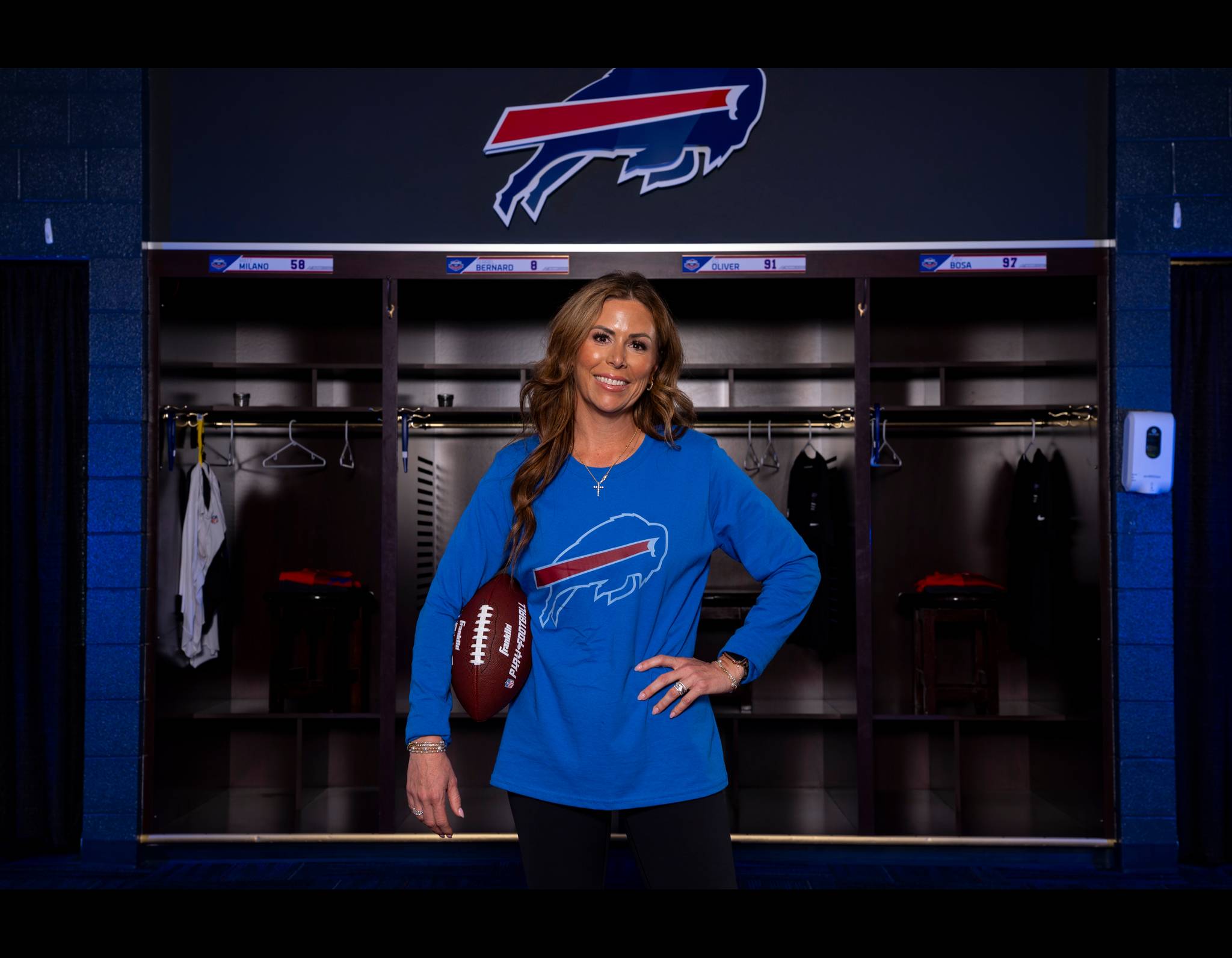 Woman in blue Bills shirt smiling in team locker room, holding football in front of player lockers with team logo above