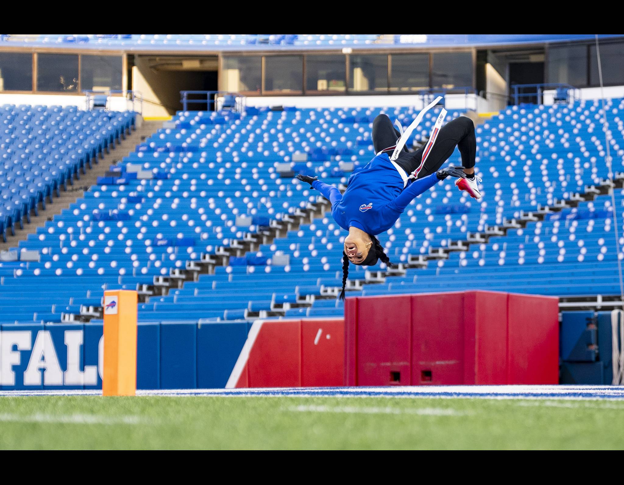 # Alt Text:

Athlete performing a backflip on a football field in an empty stadium with blue seats, yellow padding, and red barriers visible in the background.