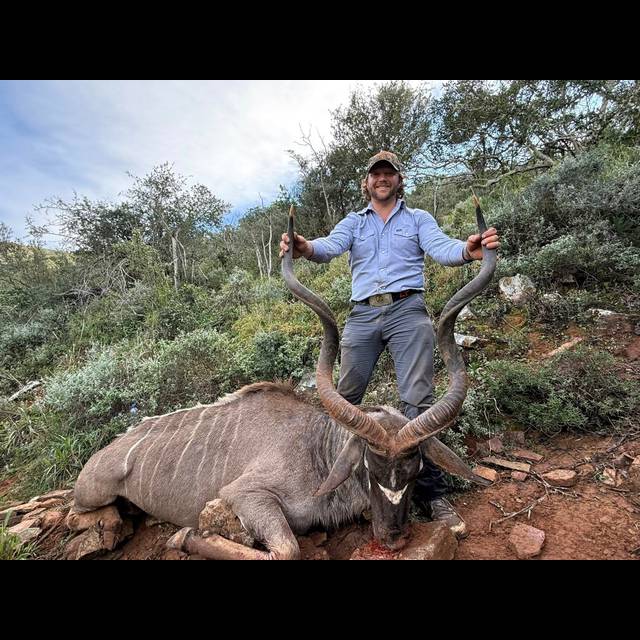 A man in a light blue shirt and cap poses behind a large antelope with impressive curved horns in a scrubland setting.