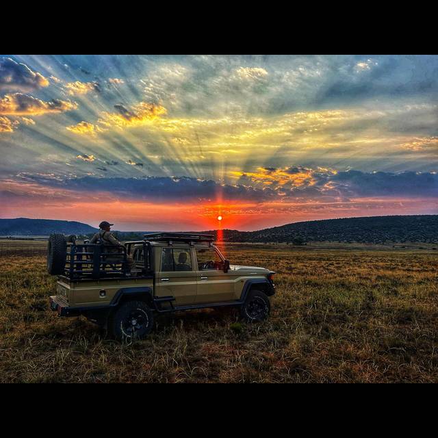 A safari vehicle with a person on top stops on an African savanna at sunset, with dramatic golden rays streaming through clouds above the horizon and rolling hills in the distance.