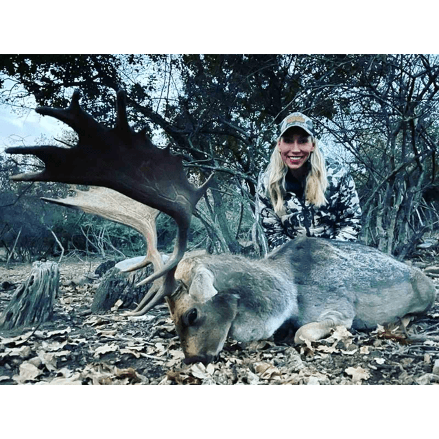 Woman in camouflage jacket and white cap posing next to a harvested deer with large antlers in a wooded outdoor setting.