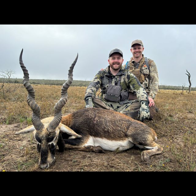 Two hunters in camouflage pose with a harvested antelope featuring long spiral horns on an open savanna landscape under an overcast sky.
