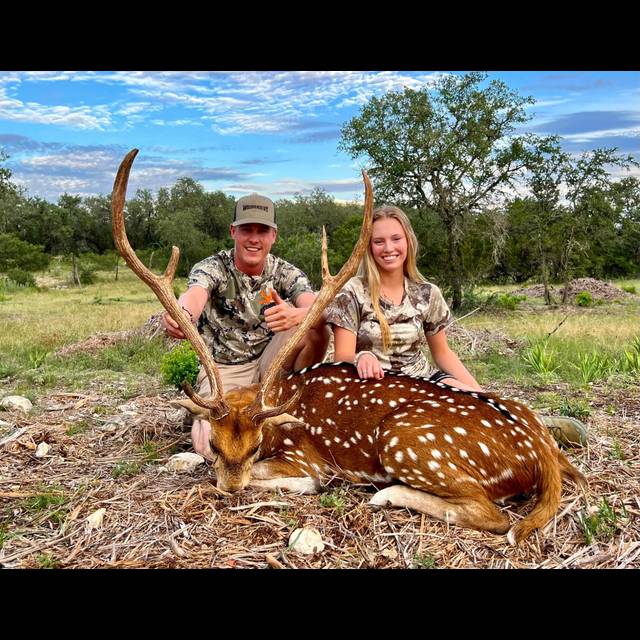 Two hunters in camouflage pose with a harvested axis deer with large antlers in a brushy, wooded landscape under a partly cloudy sky.