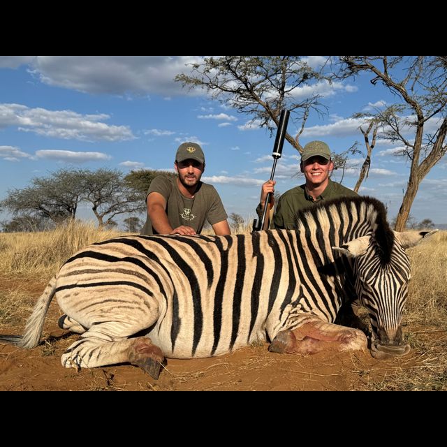 Two hunters pose with a harvested zebra in an African savanna landscape, with dry grass, scattered acacia trees, and blue sky with clouds in the background.