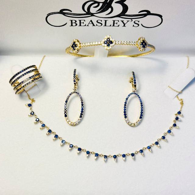 Beasley's jewelry collection featuring gold bracelets, bangles, drop earrings, and a necklace adorned with blue sapphires and diamonds displayed on white velvet.