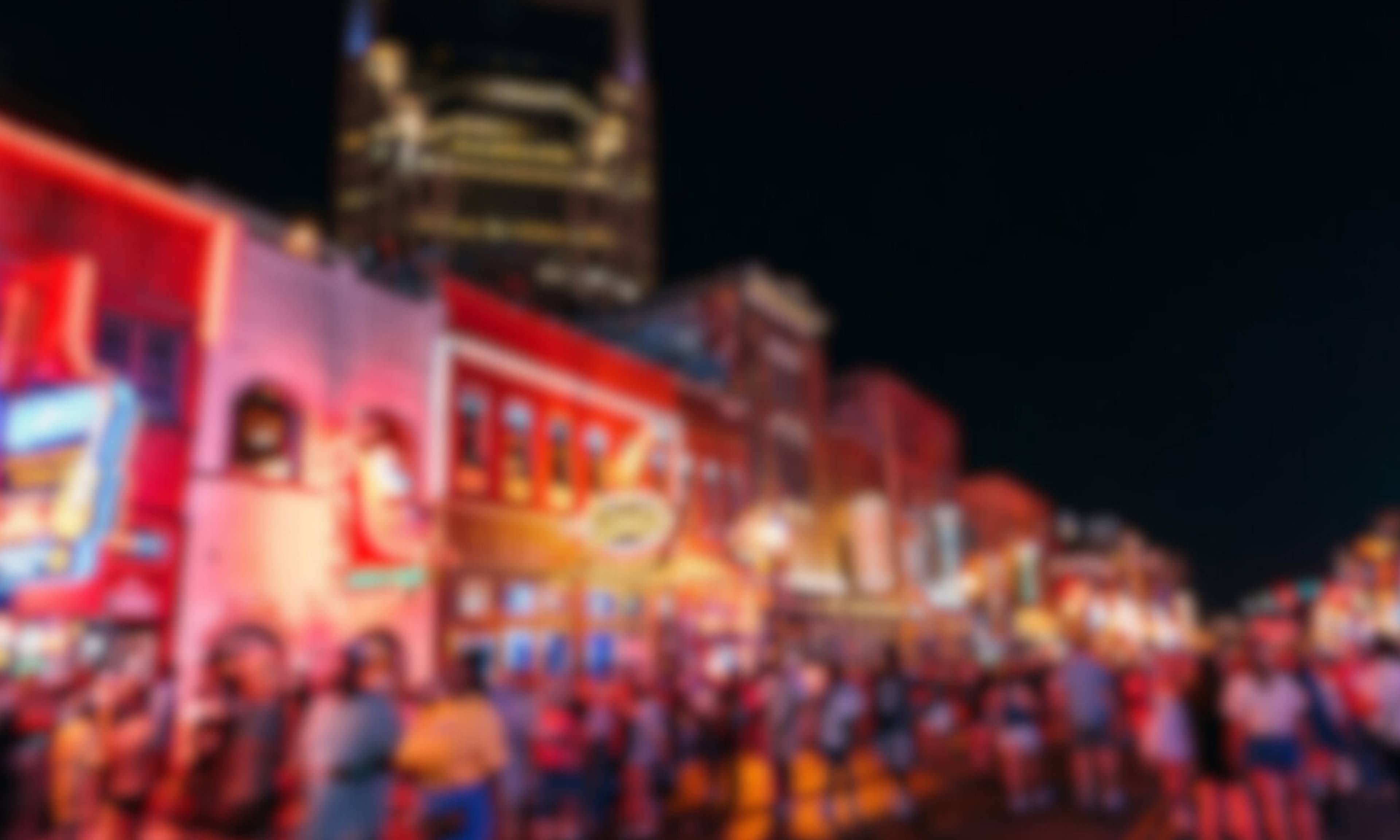 A nighttime cityscape featuring brightly illuminated red and pink buildings with neon signage, a tall illuminated tower in the background, and crowds of people gathered in the foreground against a dark sky.