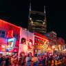 Vibrant nighttime scene of Nashville's Broadway with neon-lit honky-tonks, crowded streets filled with pedestrians, and the illuminated Nashville skyline with tall buildings in the background.