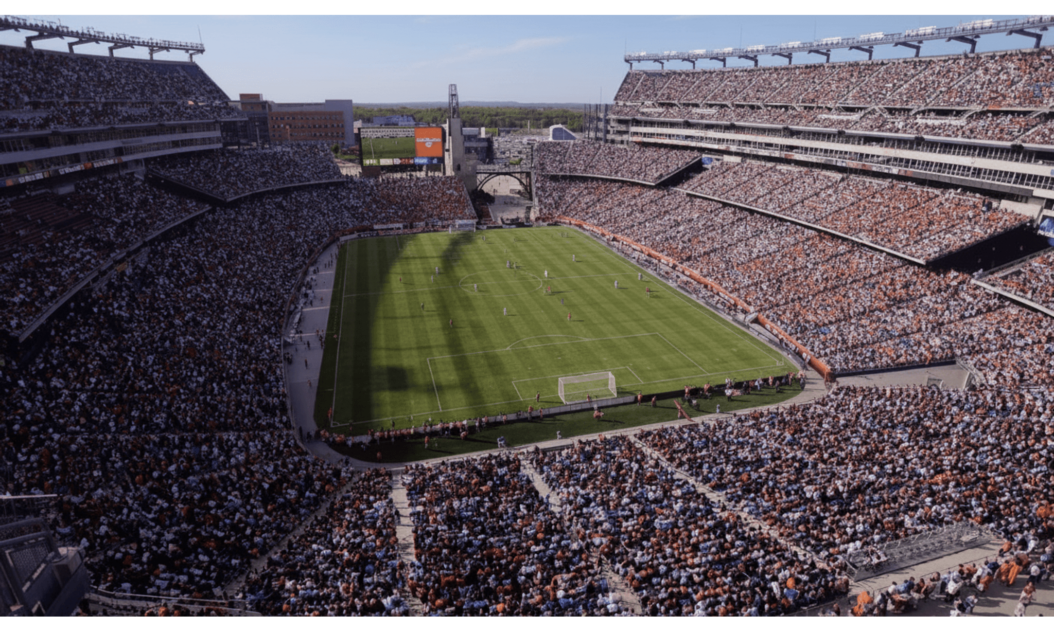 A massive football stadium packed with thousands of spectators surrounding a bright green soccer field with goal posts, large display screens, and metal bleachers under clear sky.