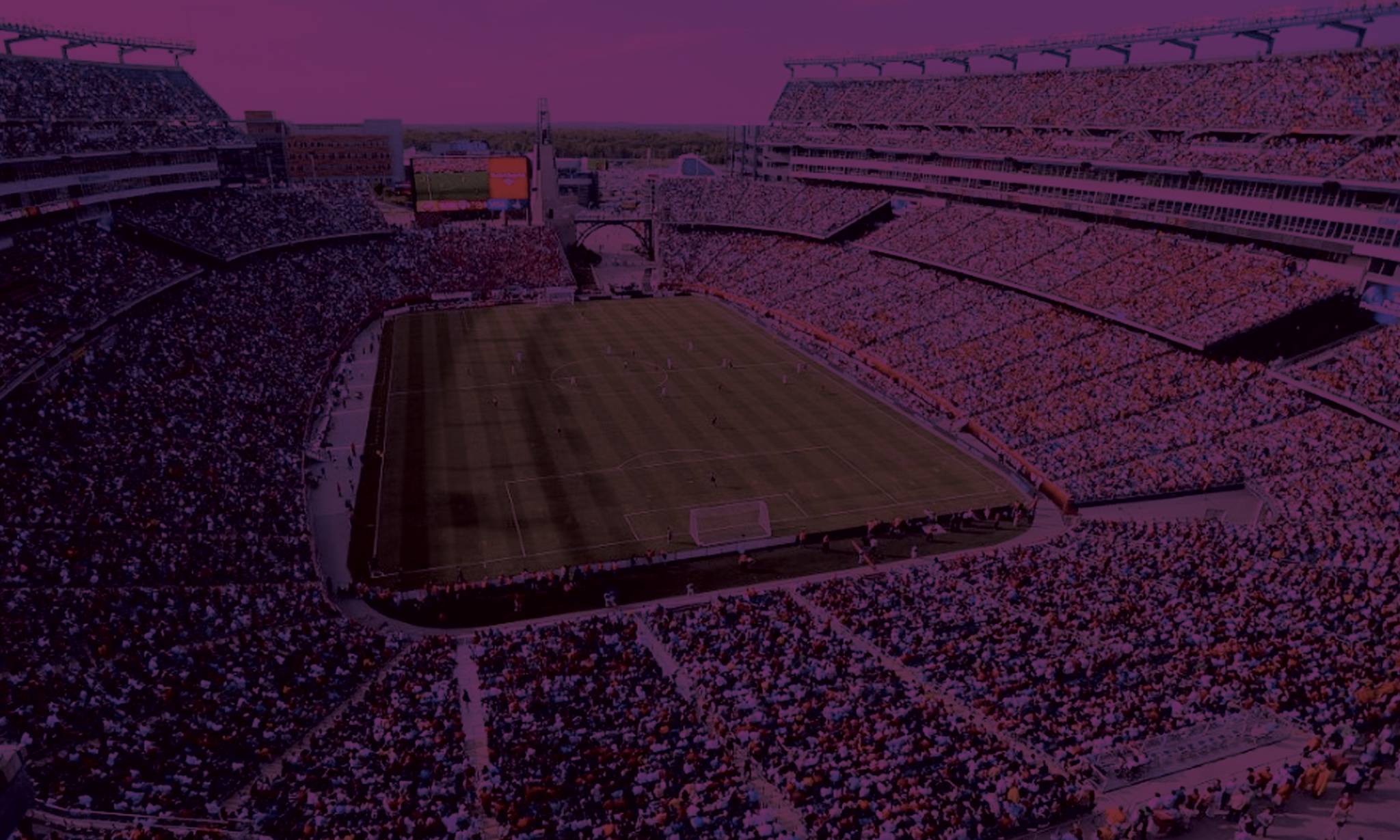 A large football stadium filled with crowds of spectators in the stands, with a grass field visible in the center under a purple-tinted sky and dramatic lighting.