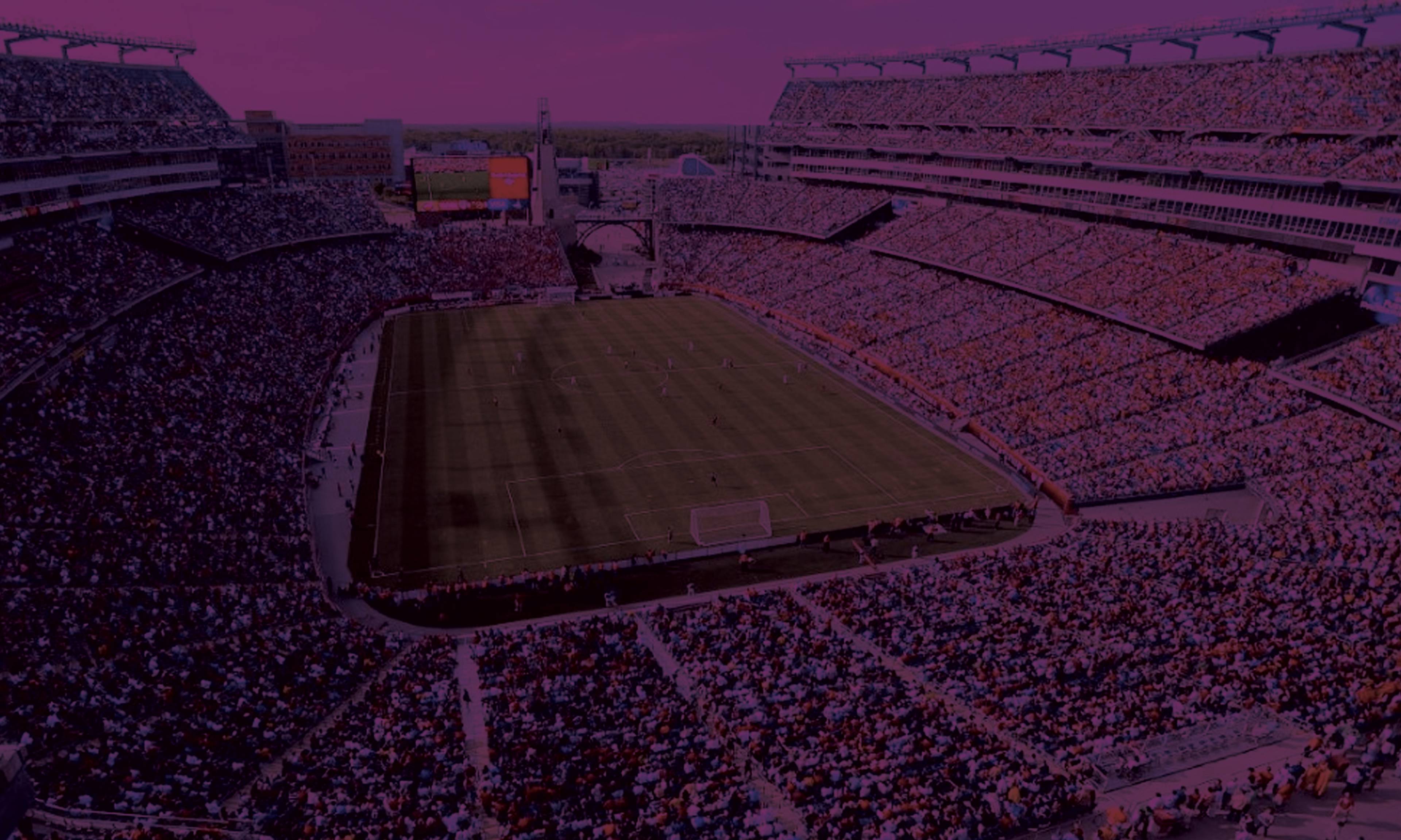A large football stadium filled with crowds of spectators in the stands, with a grass field visible in the center under a purple-tinted sky and dramatic lighting.