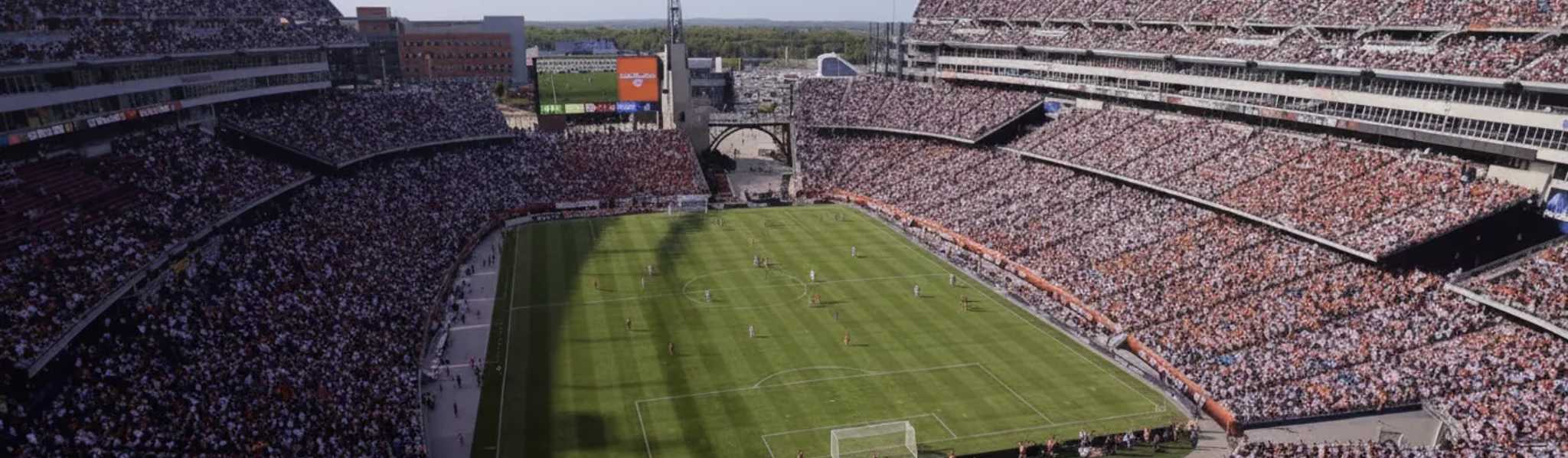 A large football stadium packed with thousands of spectators watching a match on a sunny day, with green pitch, scoreboard, and surrounding stands filled with fans.