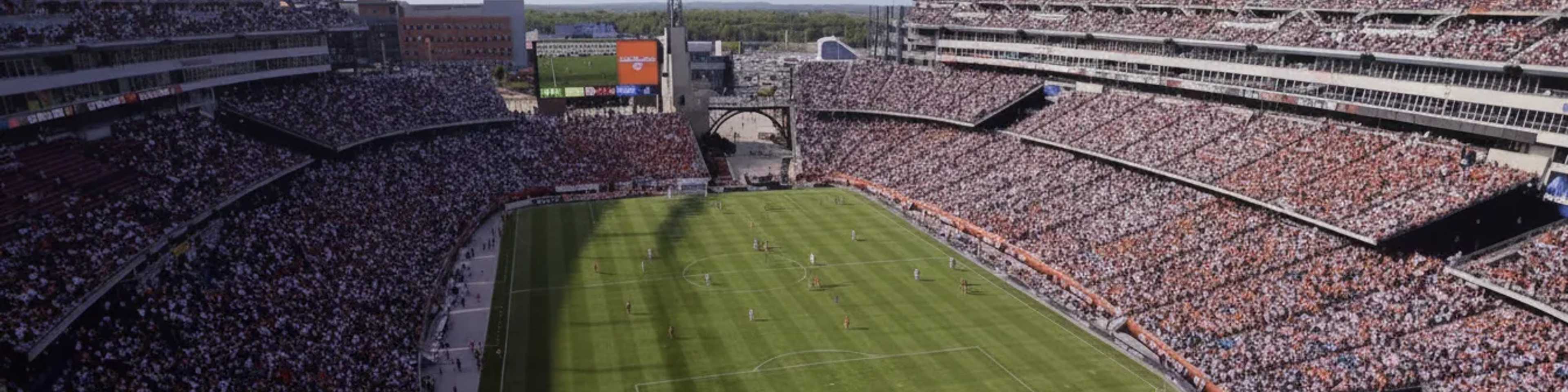 A large football stadium packed with thousands of spectators watching a match on a sunny day, with green pitch, scoreboard, and surrounding stands filled with fans.