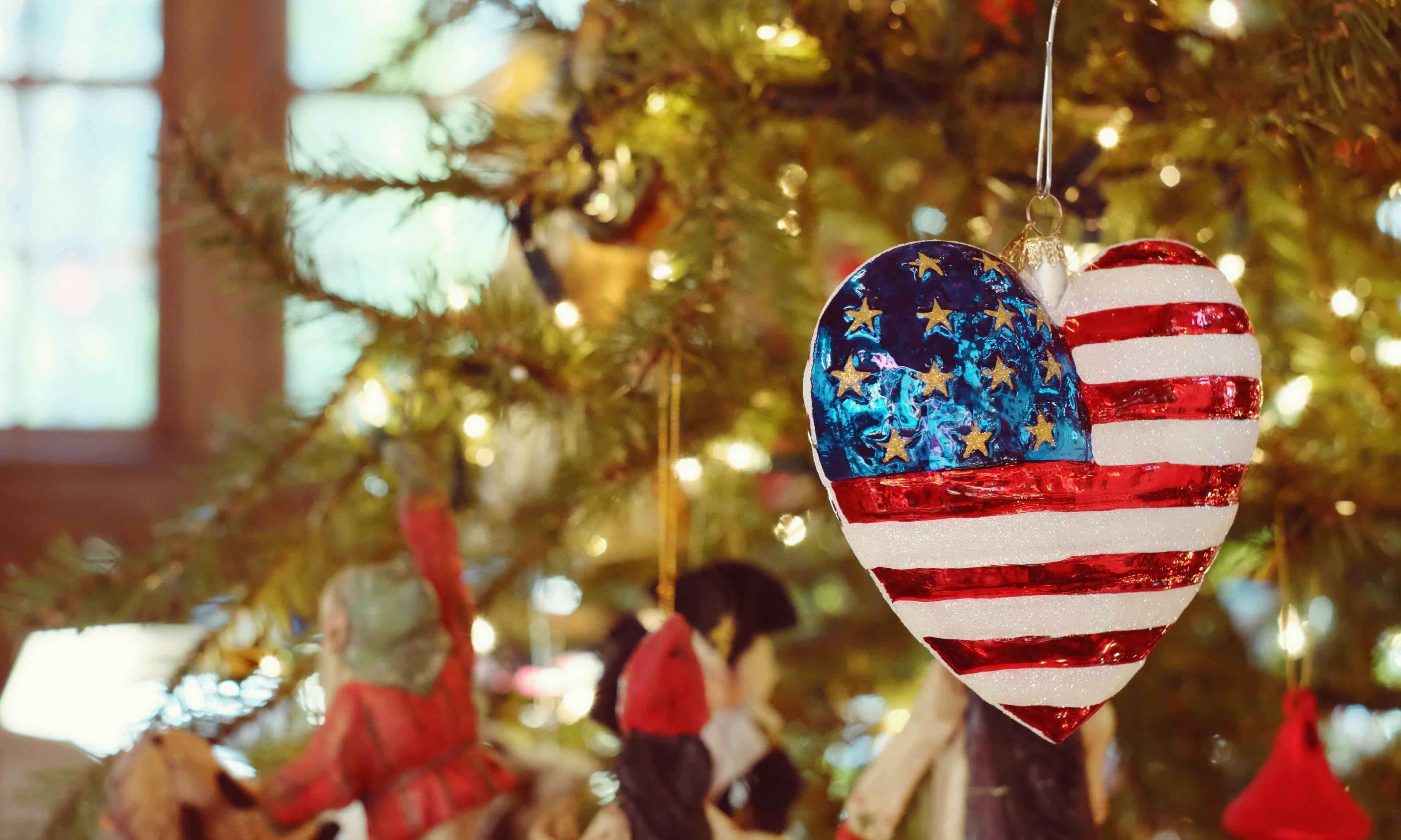 An American flag-themed heart-shaped ornament hangs on a decorated Christmas tree with glowing lights and other festive ornaments in the background.