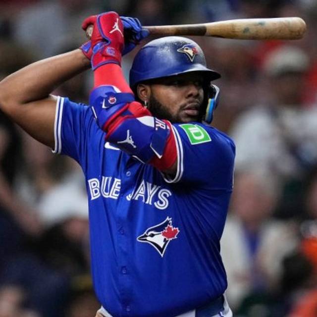 A Toronto Blue Jays baseball player in blue uniform holds a bat over his shoulder, wearing a batting helmet and red gloves, ready to bat during a game.