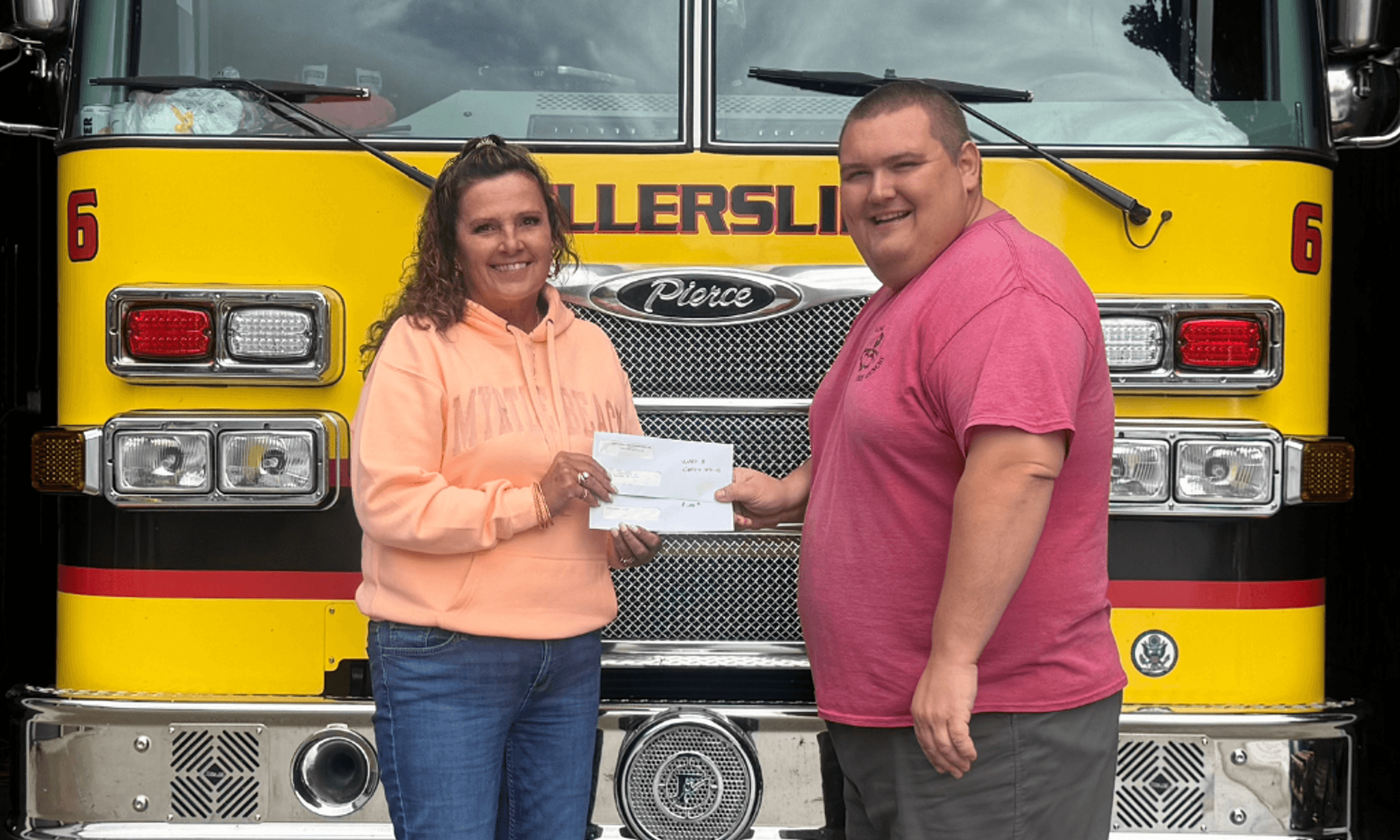 A woman in a peach hoodie and a man in a pink shirt stand in front of a yellow fire truck, both smiling while holding a check.