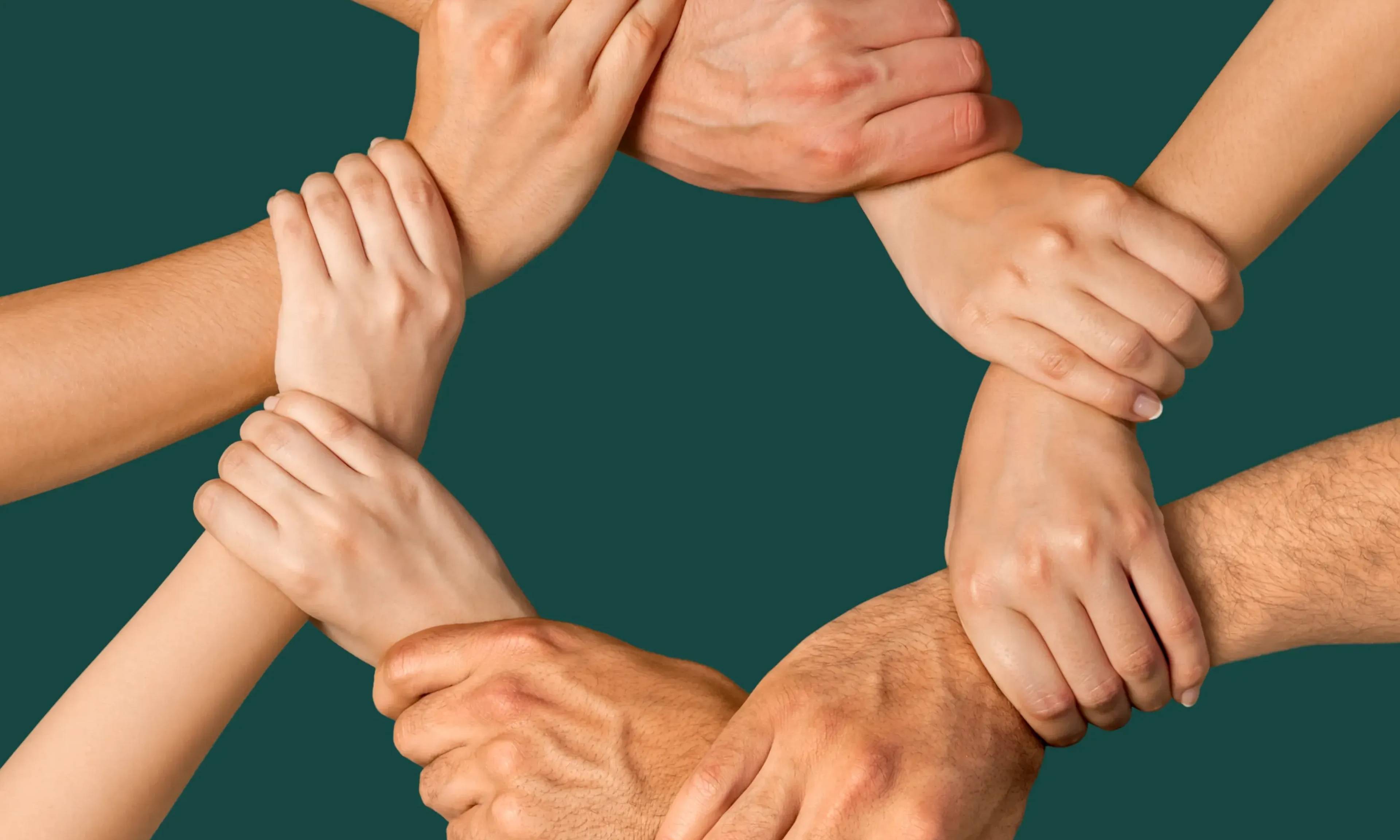 Multiple hands joined together in a circle against a teal background, symbolizing unity, teamwork, and collaboration among diverse people.
