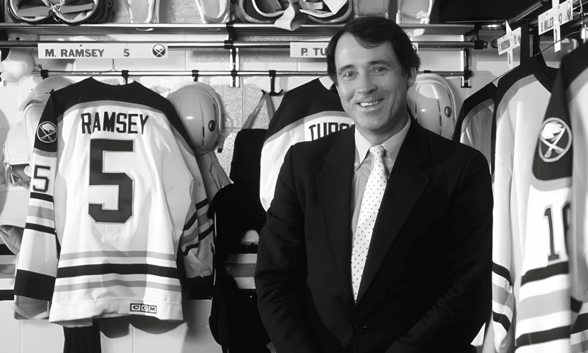 Black and white photograph of a man in a business suit smiling in a hockey locker room, standing beside jersey number 5 with "RAMSEY" on it.