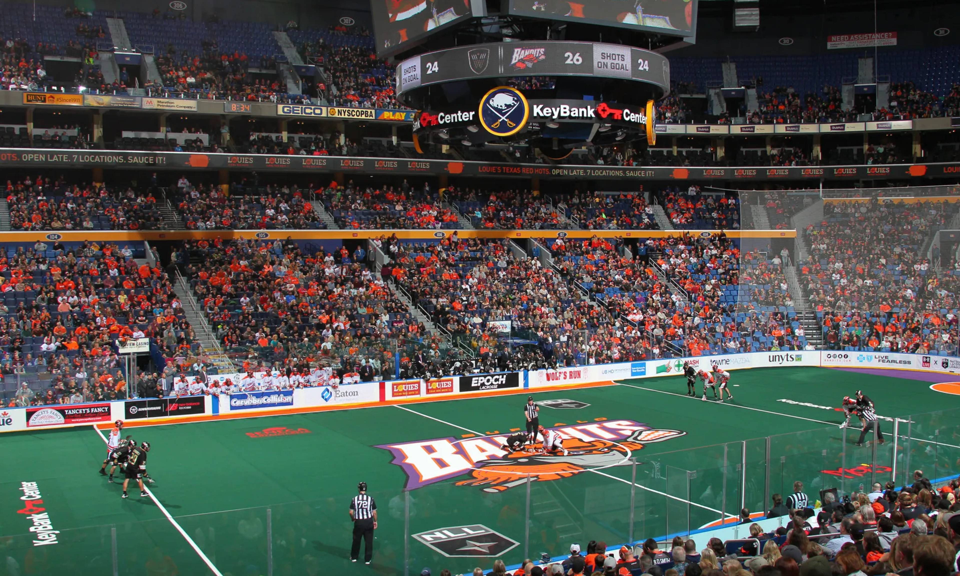 An indoor lacrosse game at KeyBank Center with teams on a green field, referees, crowded stands filled with fans in orange and blue, and large scoreboard displays overhead.