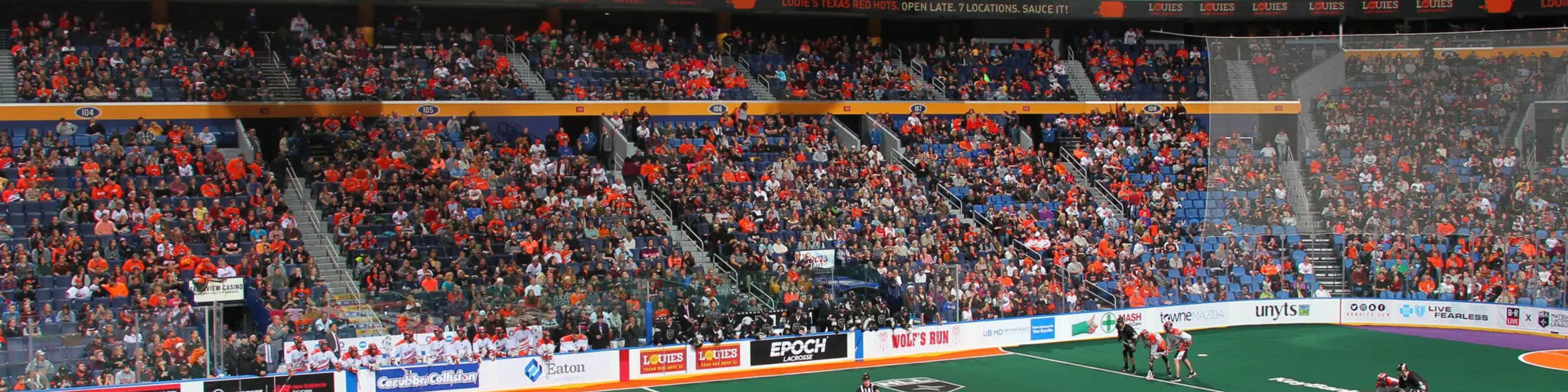 Indoor lacrosse game at KeyBank Center with Buffalo Sabres branding, packed stadium with orange and blue team uniforms, players on green field, scoreboard showing 24-26, referees on court.
