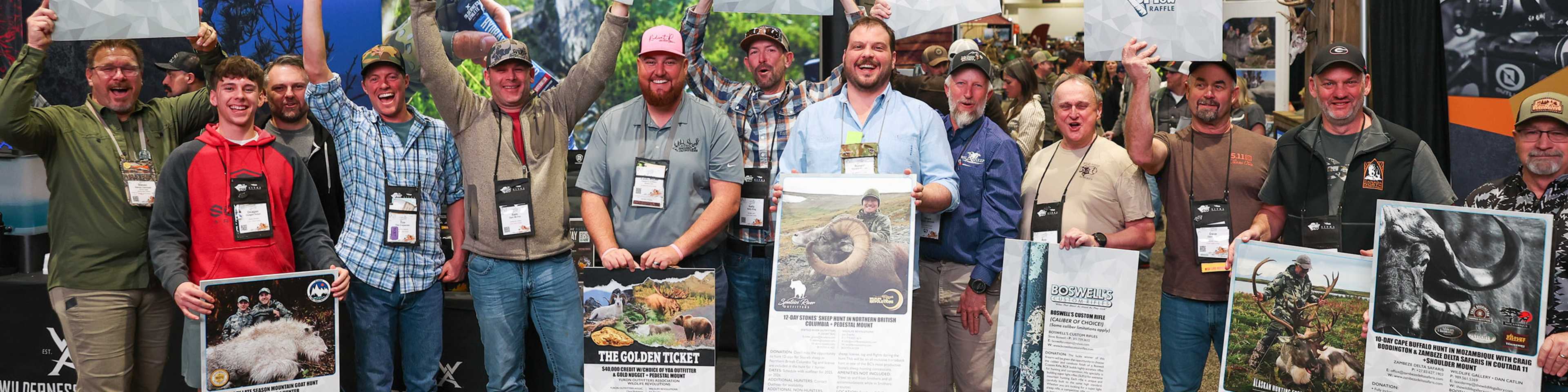 A group of hunters and outdoor enthusiasts posing together at an indoor convention or expo, holding large informational posters about hunting expeditions and wildlife, with mounted animal displays visible in the background.