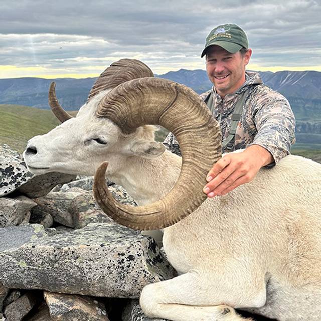 Hunter poses with a harvested Dall sheep featuring curved horns, sitting on rocky terrain with mountains visible in the background under a cloudy sky.