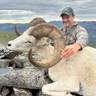 Hunter poses with a harvested Dall sheep featuring curved horns, sitting on rocky terrain with mountains visible in the background under a cloudy sky.