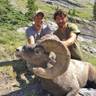 Two men posing with a bighorn sheep in a mountainous landscape with rocky terrain and green vegetation in the background.