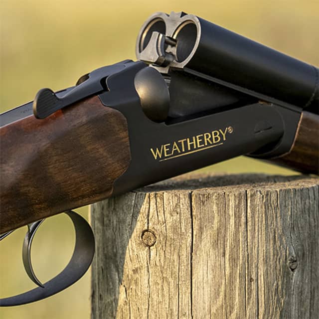 A Weatherby shotgun with a dark barrel and wood furniture rests on a wooden post against a blurred yellow-green background.