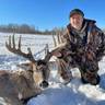 Hunter in camouflage jacket posing with a large-antlered deer in snow-covered landscape under clear blue sky.