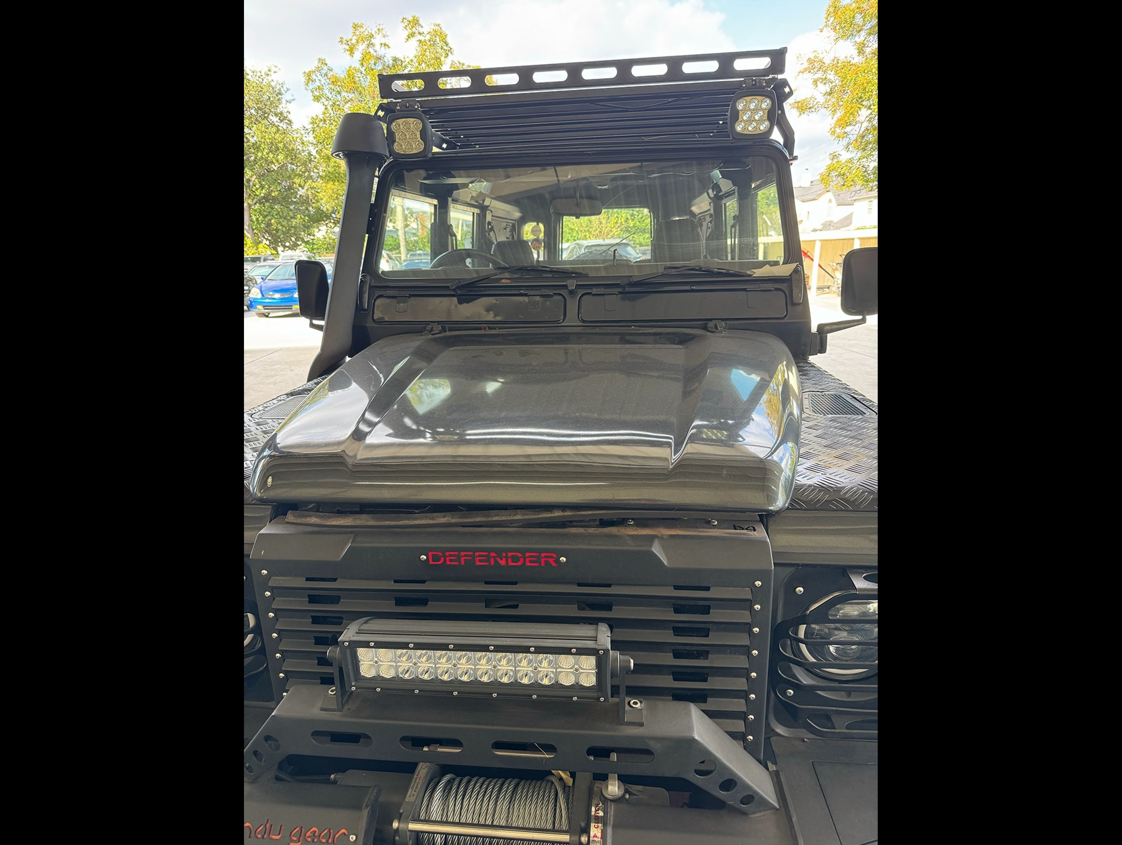 A black Land Rover Defender with custom modifications including LED lights on the roof rack and front bumper, parked in a dealership lot on a sunny day with trees in the background.