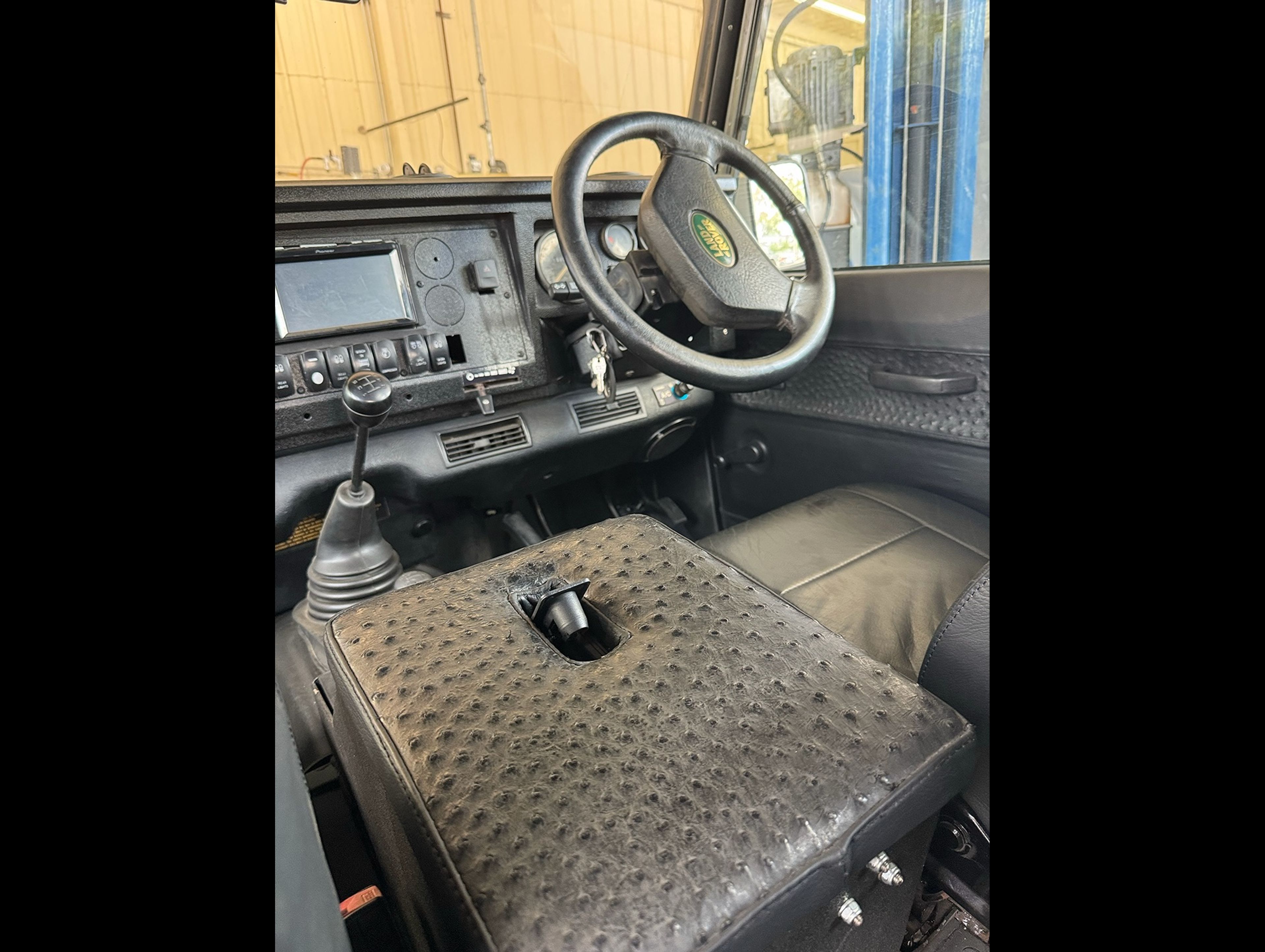 Interior of a Land Rover vehicle showing the steering wheel, dashboard with controls, and a textured seat with a cup holder, viewed from the passenger side.