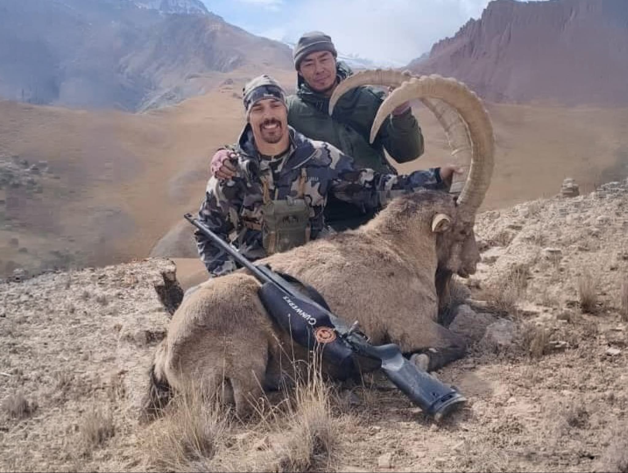 Two hunters pose with a harvested argali sheep in a mountainous valley landscape, displaying the large curved horns characteristic of the species.