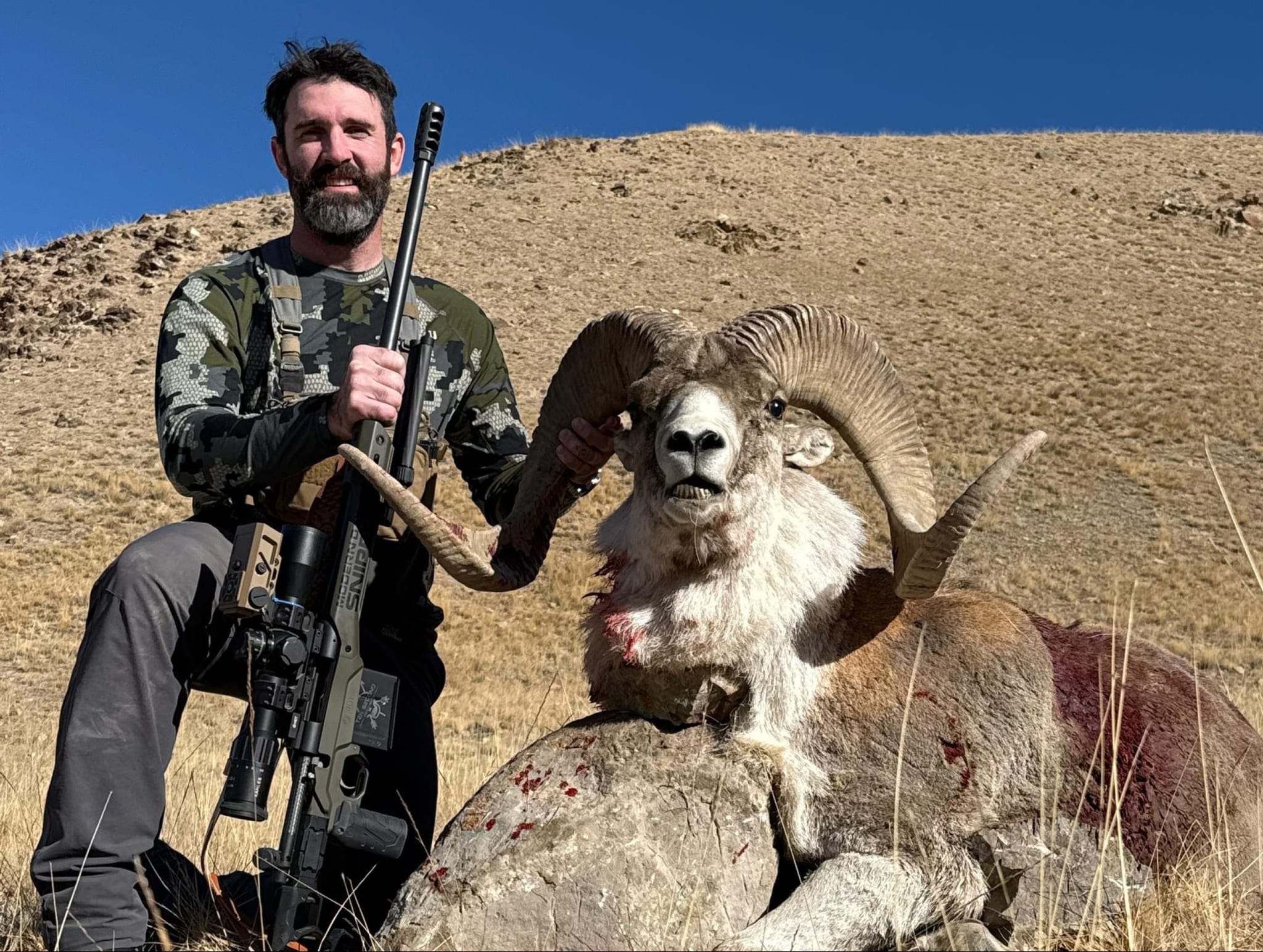 Hunter posing with a rifle next to a harvested bighorn sheep with large curved horns on a desert hillside under clear blue sky.