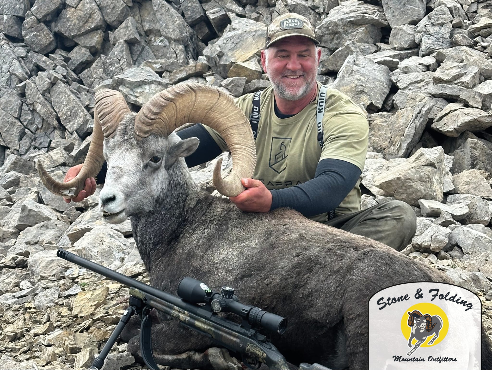 Hunter posing with a harvested bighorn sheep with curved horns next to a rifle with scope, rocky terrain in background.