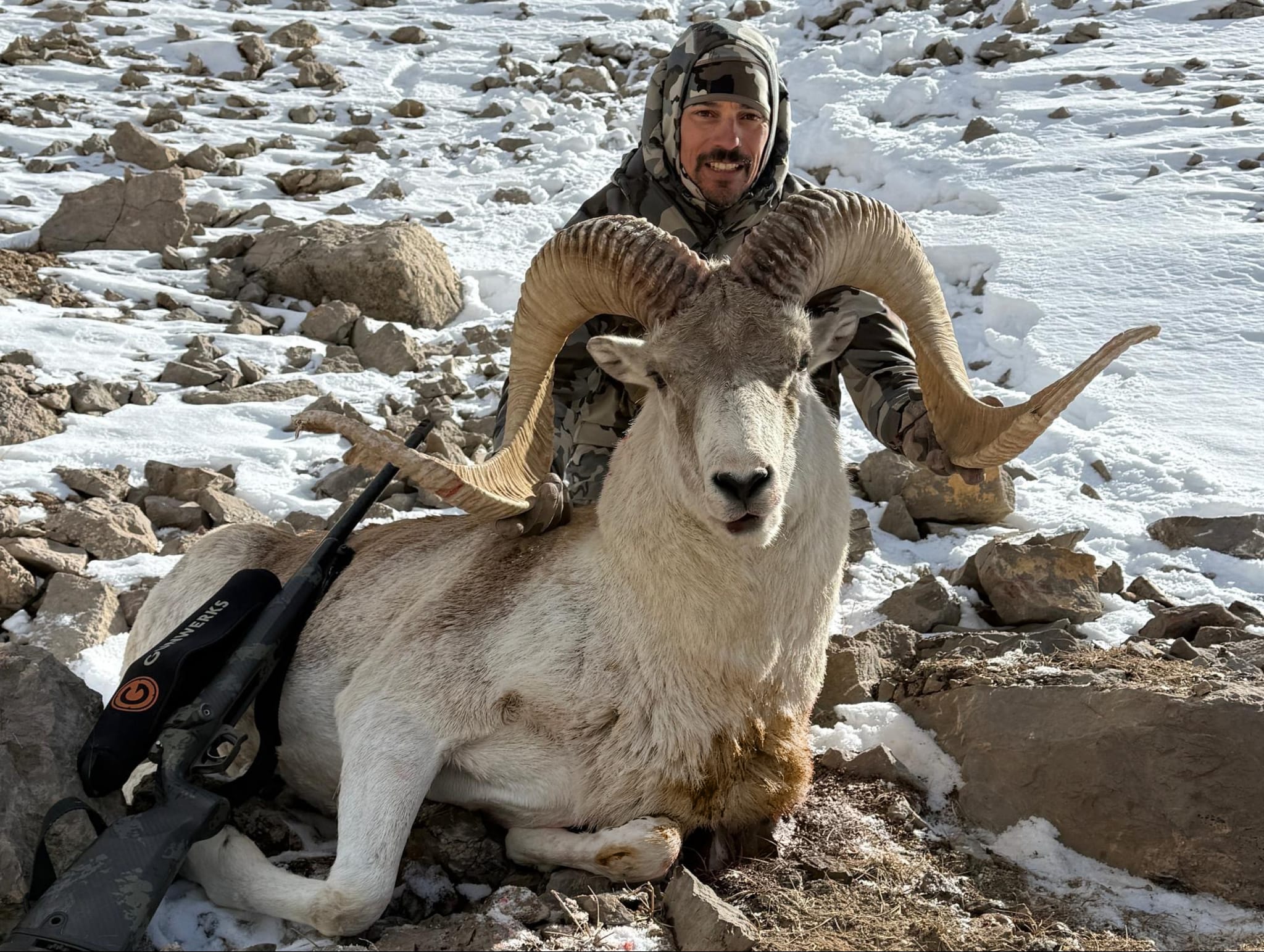 Hunter posing with a harvested Dall sheep with large curved horns in a snowy, rocky mountain landscape.