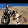 Hunter posing with a harvested bighorn sheep on an arid hillside, holding a rifle with scope, under clear blue sky.
