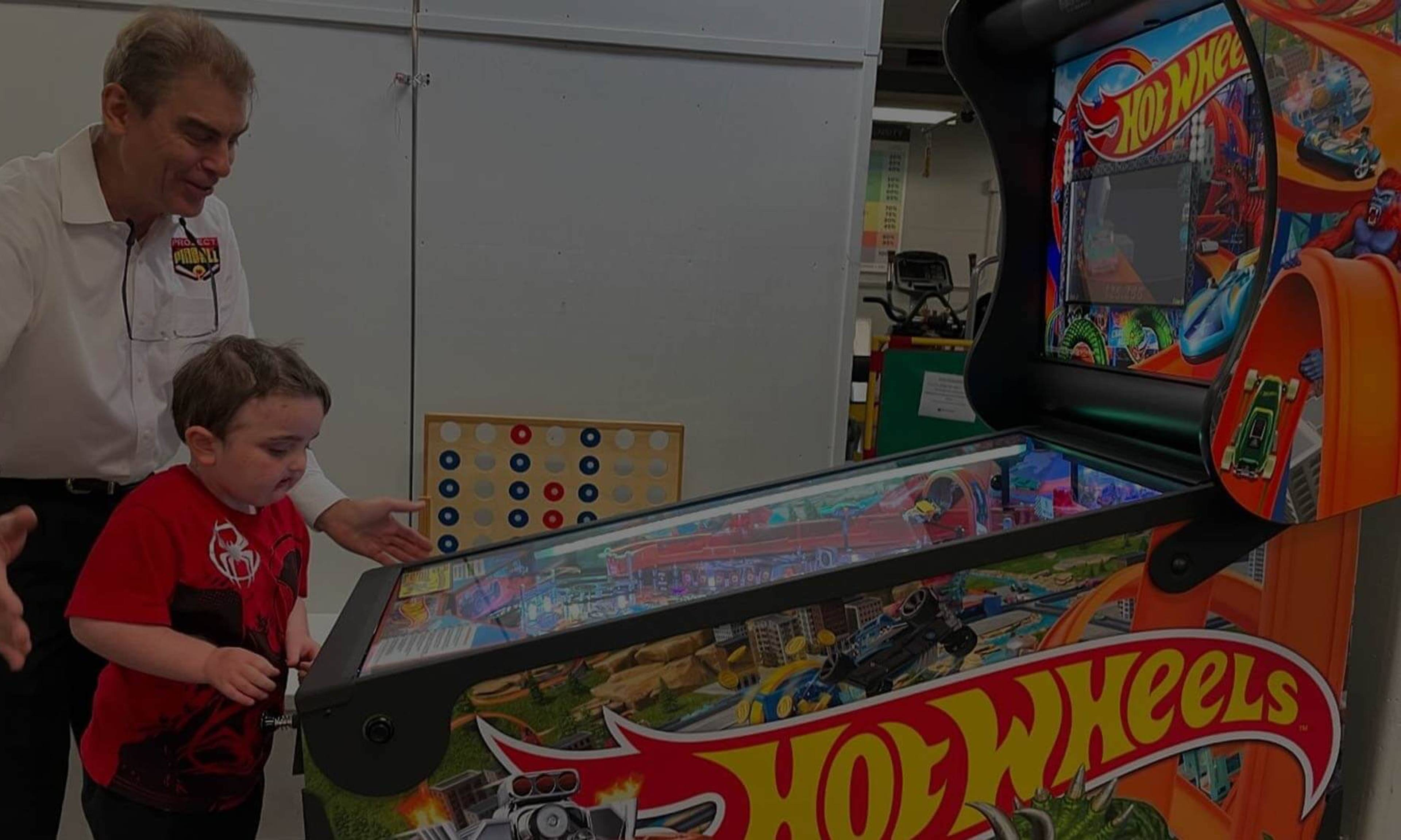 Adult and child playing Hot Wheels pinball machine, with colorful game board and Connect Four game in background.