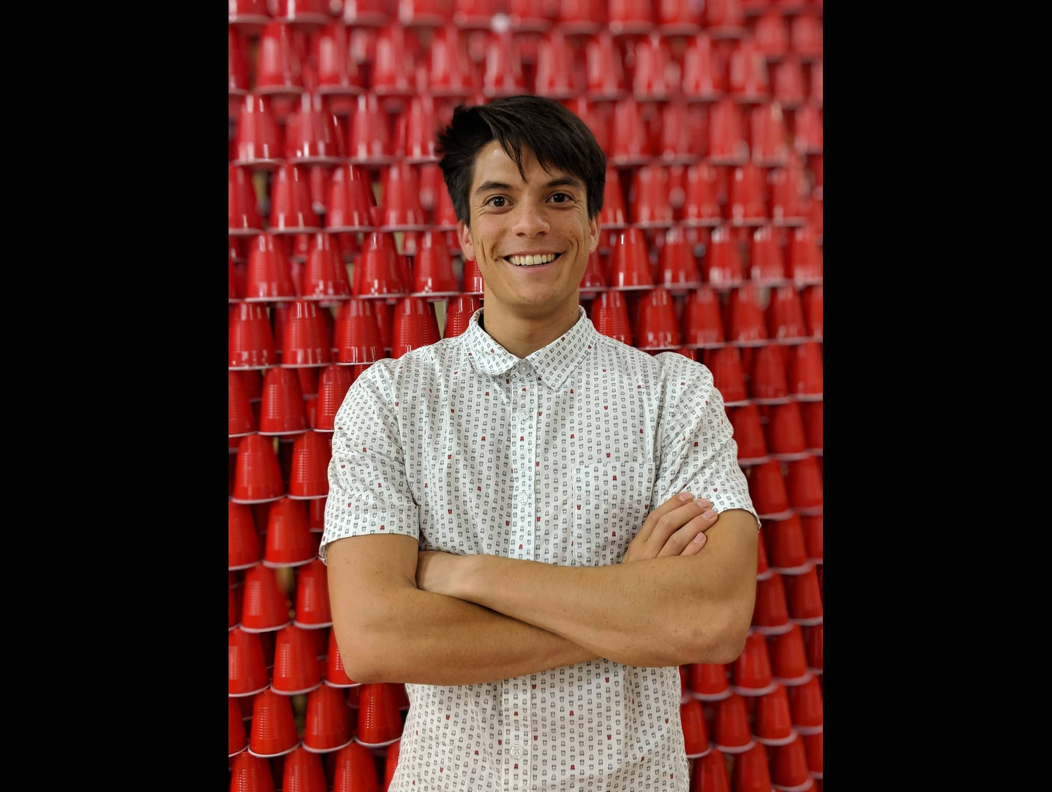 Smiling person in patterned shirt standing in front of a wall of red plastic cups with arms crossed