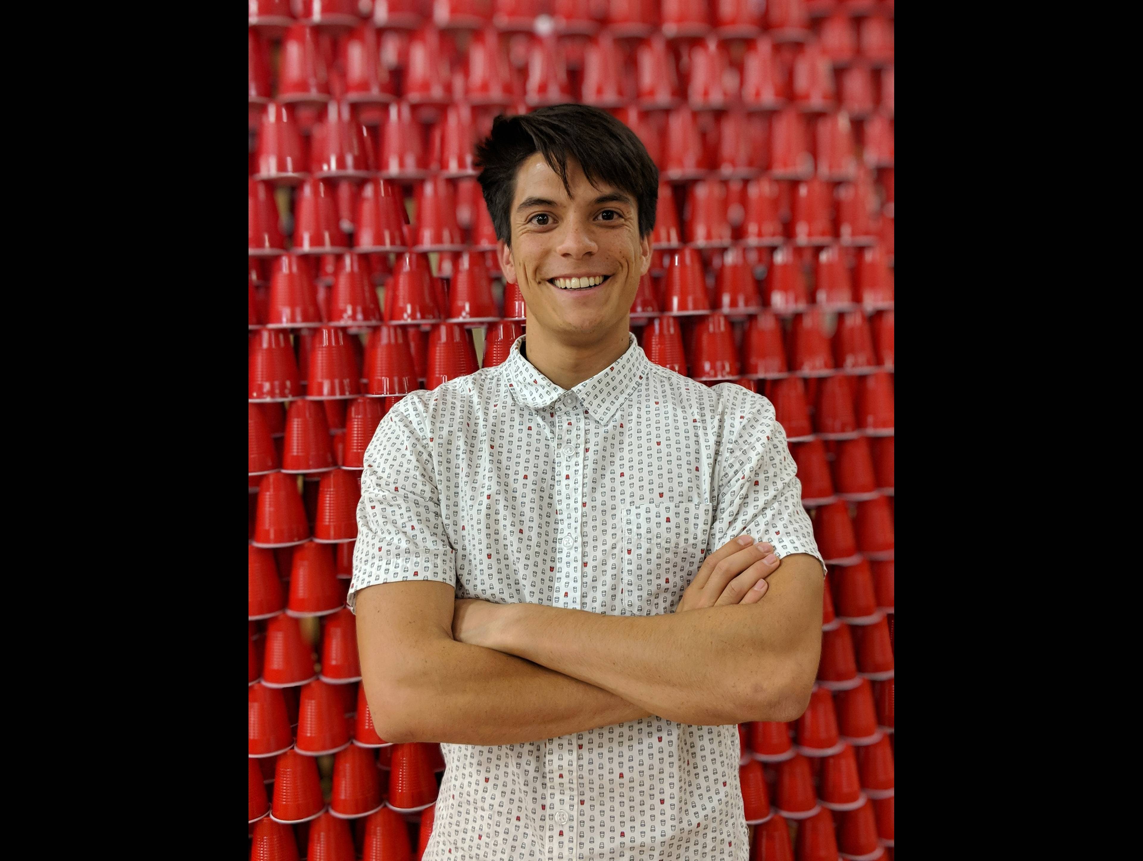 Smiling person in patterned shirt standing in front of a wall of red plastic cups with arms crossed