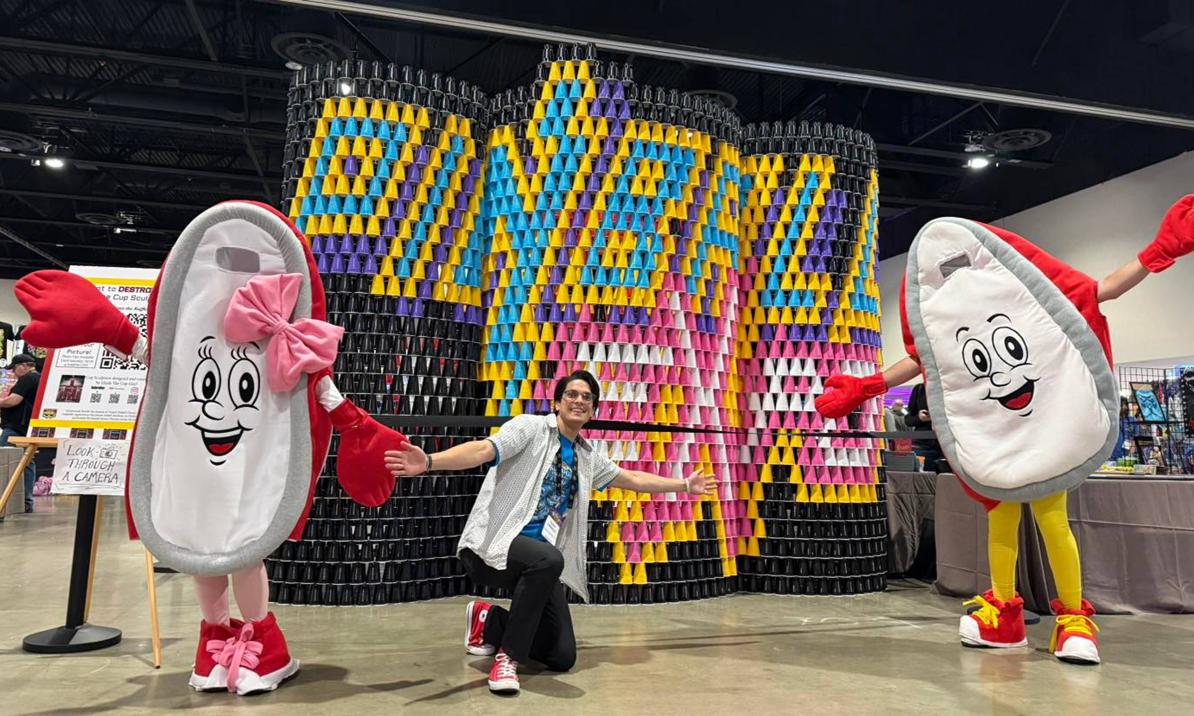Two cute mascot characters flank a person posing in front of a large colorful cup display at a convention.