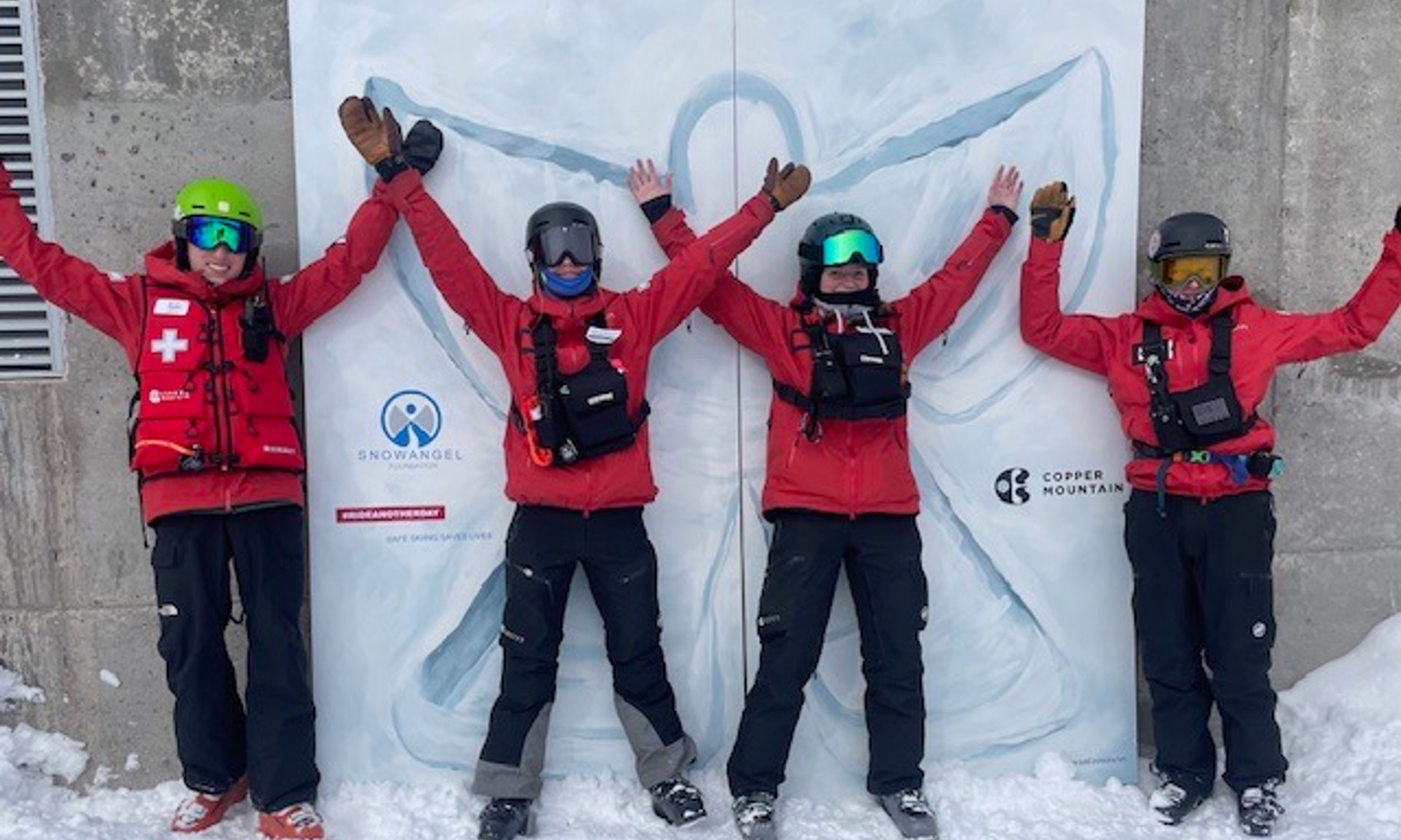 Four skiers in red jackets and black pants stand with raised arms in front of a snowy mountain backdrop and white banner with logos.