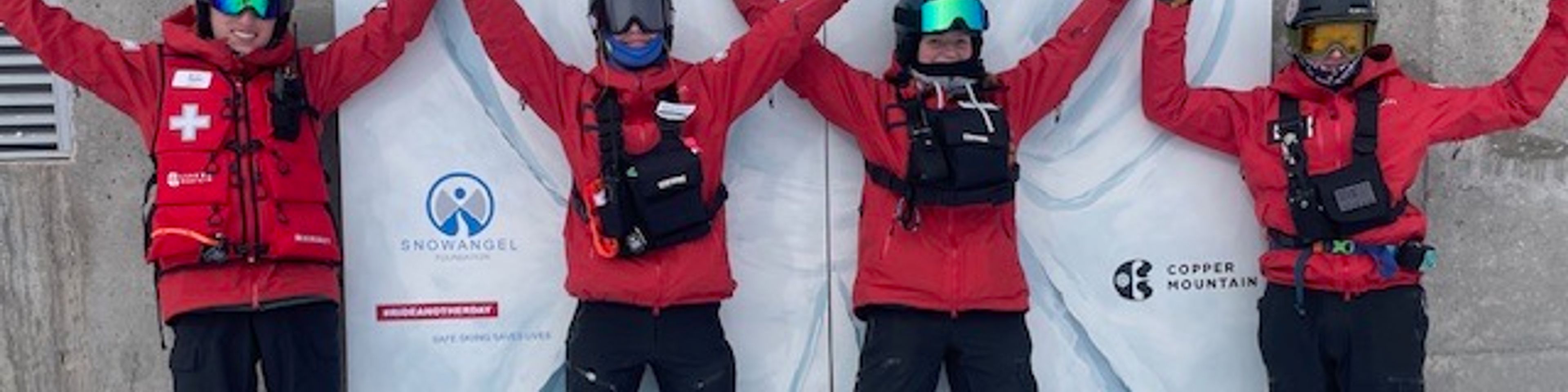 Four skiers in red jackets and winter gear stand in the snow with their arms raised in celebration in front of a white banner with logos.