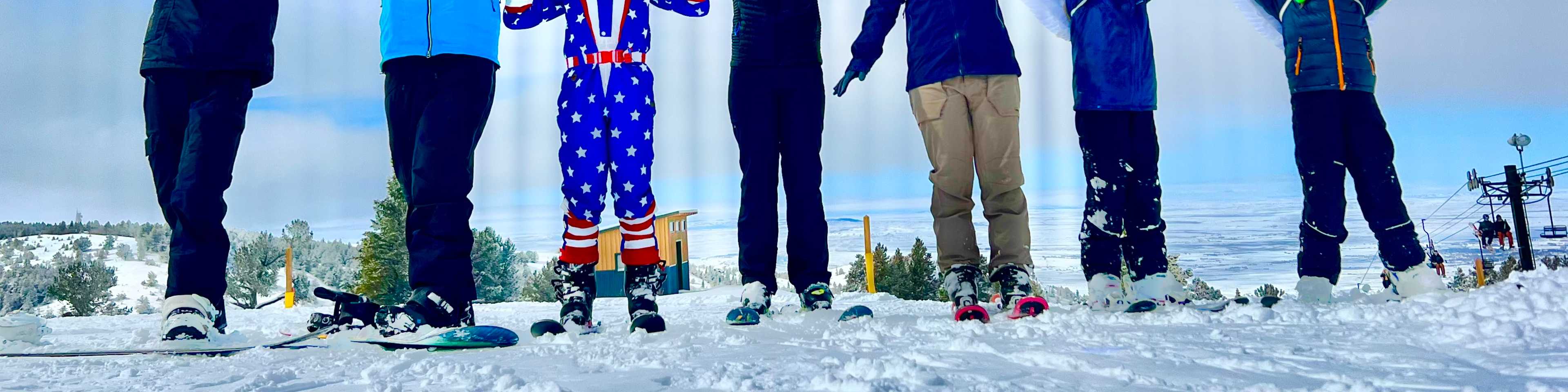 Group of skiers and snowboarders celebrating with arms raised on a snowy mountain slope under blue sky, wearing colorful winter gear and goggles.