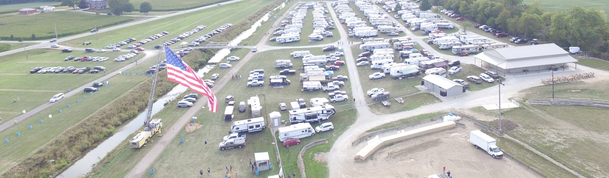 Aerial view of a large RV and camper storage facility with organized rows of white trailers, an American flag, and surrounding rural farmland with fields and trees.