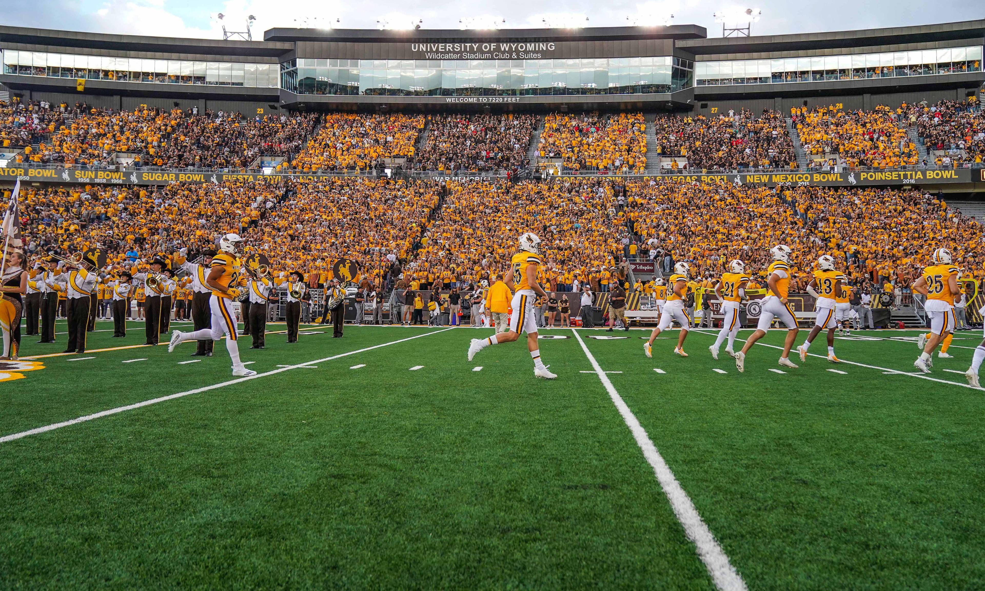 University of Wyoming football team in yellow uniforms warming up on the field at a packed stadium with thousands of fans in the stands.